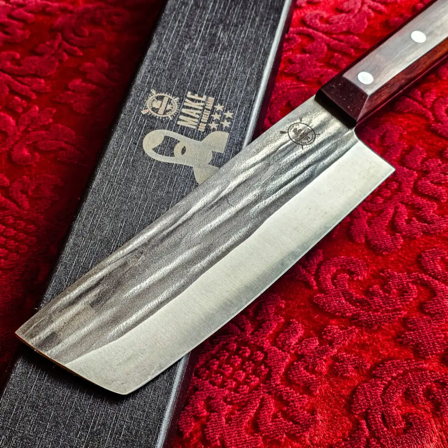 Nakiri Kockkniv Red Velvet #15 vacker kniv från BBQmonsters serie av Asiatiska kockknivar - liknande kniv säljs för det tredubbla på andra sidor! Söker du en serie av välarbetade och lättskötta kockknivar? Ta en kik på BBQmonsters egna serie knivar! Samtliga knivar i serien kommer i en svart presentförpackning med BBQmonsters sigill "Make BBQ great again" tryckt på locket. Knivarna från BBQmonster har även BBQmonsters logo ingraverad på bladet. Vi sätter bara vår logo på prylar som vi verkligen tycker om - detta är en sådan. Introduktionspris - passa på! En perfekt till grillnörden, en present till pappa på farsdag eller till dig själv helt enkelt! Nakiri kockkniven i serien Red velvet har ett abrupt avslut av bladet, nästan likt en avbruten kniv. Bladets höjd är sett till bladet längd relativt högt. Detta gör att du här har en lättstyrd kniv som du med enkelhet kan hacka upp stora mängder grönsaker i symmetriska former  . Kniven har en längd som gör den enkel att kontrollera om du önskar använda den för att dekorera en frukt eller grönsak med ett snyggt inristat mönster. En bra allround grönsakskniv som ersätter en klassisk "petty-kniv". Bladet på denna Red Velvet #15 är vackert dekorerat med mjuka följsamma mönster inhamrat i stålet. Bladet har en perfekt balans mellan smidigt och robust. Eggen är extremt vass och lättskött. Varje blad är unikt i sitt utseende vilket tillsammans allts som allt gör detta kniven rejält "intstagramvänlig" och photogenique. Bladet är genomgående och avslutas med det snygga fyrkantiga handtaget som är av rött ädelträ och som har tre nitar av mässing. Skaftet ger ett skönt grepp och är enkelt att rengöra. Handtaget passar både höger- och vänsterhänta. Kolstål med blandning X50CrMoV15 Hårdhet 56° HRC vilket är ett relativt mjukt stål - något som gör att du själv enkelt kan hålla din kniv vass. Total längd: 31 cm Längd på bladet: 18 cm Längd på skaftet: 13cm Vikt: 276 gram