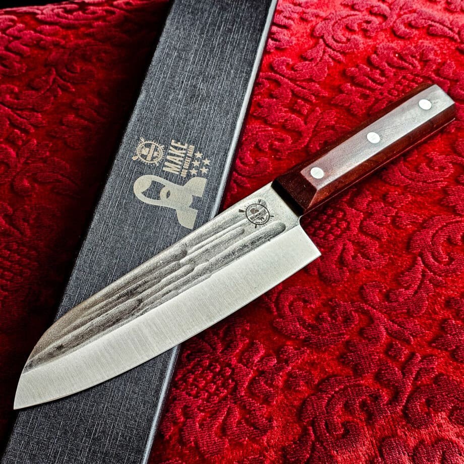 Santoku kockkniv Red Velvet #16 vacker kockkniv från BBQmonsters serie av Asiatiska kockknivar - liknande kniv säljs för det tredubbla på andra sidor! Söker du en serie av välarbetade och lättskötta kockknivar? Ta en kik på BBQmonsters egna serie knivar! Samtliga knivar i serien kommer i en svart presentförpackning med BBQmonsters sigill "Make BBQ great again" tryckt på locket. Knivarna från BBQmonster har även BBQmonsters logo ingraverad på bladet. Vi sätter bara vår logo på prylar som vi verkligen tycker om - detta är en sådan. Introduktionspris - passa på! En perfekt till grillnörden, en present till pappa på farsdag eller till dig själv helt enkelt! Santoku betyder "tre syften" på japanska och det berättar att detta är en kockkniv för både fisk, kött och grönsaker. Det lite kortare bladet gör att det är enkelt att arbeta med precision. Behöver du ett längre blad för att tranchera större stekar så ta en kik på Gyuto varianten i denna Red velvet serie. Bladet på denna Red Velvet #16 är vackert dekorerat med mjuka följsamma mönster inhamrat i stålet. Bladet har en perfekt balans mellan smidigt och robust. Eggen är extremt vass och lättskött. Varje blad är unikt i sitt utseende vilket tillsammans allts som allt gör detta kniven rejält "intstagramvänlig" och photogenique. Bladet är genomgående och avslutas med det snygga fyrkantiga handtaget som är av rött ädelträ och som har tre nitar av mässing. Skaftet ger ett skönt grepp och är enkelt att rengöra. Handtaget passar både höger- och vänsterhänta. Kolstål med blandning X50CrMoV15 Hårdhet 56° HRC vilket är ett relativt mjukt stål - något som gör att du själv enkelt kan hålla din kniv vass. Total längd: 30,5 cm Längd på bladet: 17,5 cm Längd på skaftet: 13cm Vikt: 234 gram